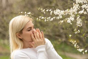 Young woman experiencing seasonal allergies and touching her nose while standing under a flowering tree